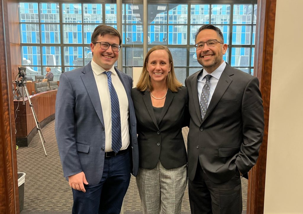 Three people in business suits stand close together and smile at the camera. 